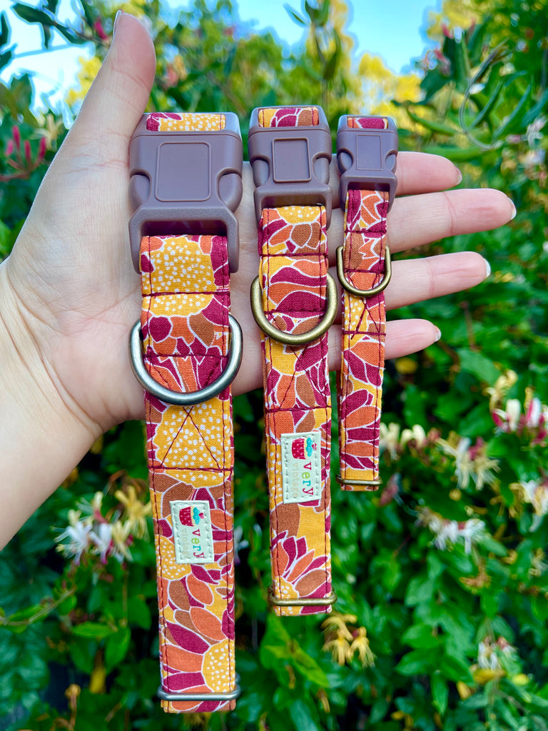 Handmade sunflower dog collar or cat collar with mustard yellow and rust red floral print, displayed outdoors in a garden setting. Made with organic cotton and antique brass or antique silver hardware.