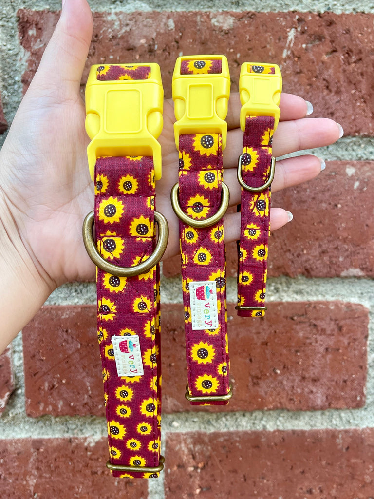 Hand holding three burgundy sunflower print dog collars with yellow buckles against brick wall background