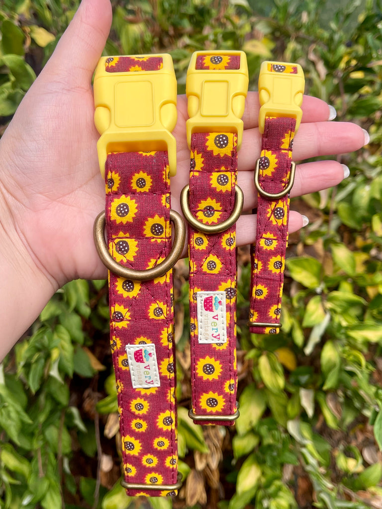 Hand holding three burgundy sunflower print dog collars with yellow buckles against green leafy background