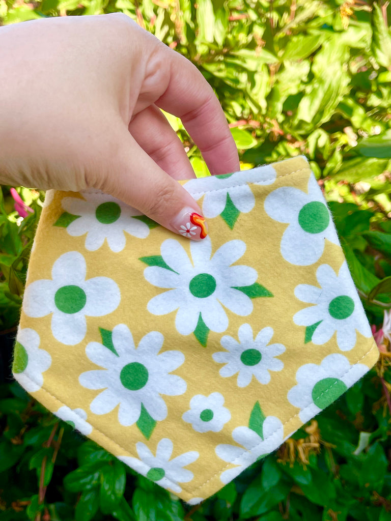 Floral dog bandana with cheerful white daisies on a butter yellow background, shown outdoors with green leaves. Lightweight and eco-friendly.