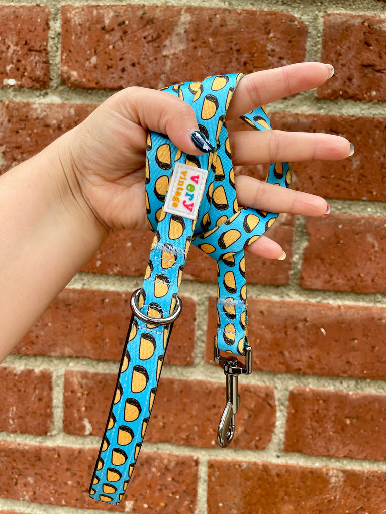 Bright taco print dog leash with black hardware held in hand in front of a red brick wall