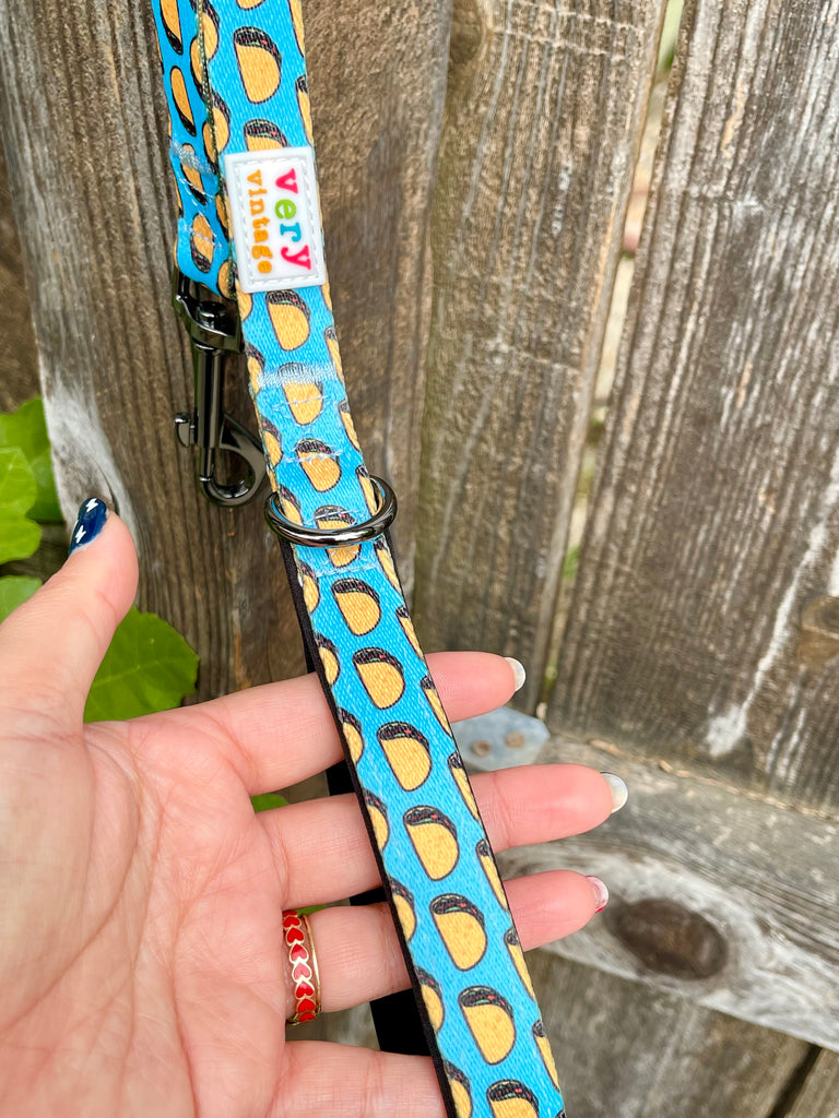 Close-up of hand holding taco print dog leash against rustic wood fence, showing metal clip and Very Vintage label