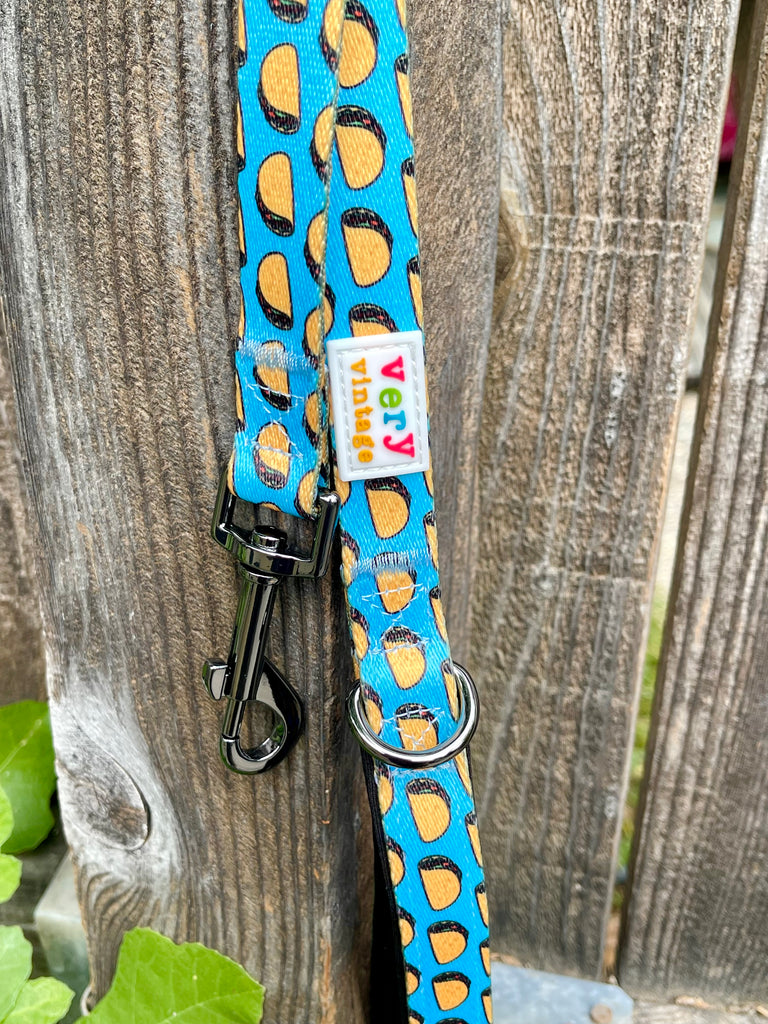 Taco-themed dog leash with black metal clip and D-ring, hanging on a wooden fence with green leaves