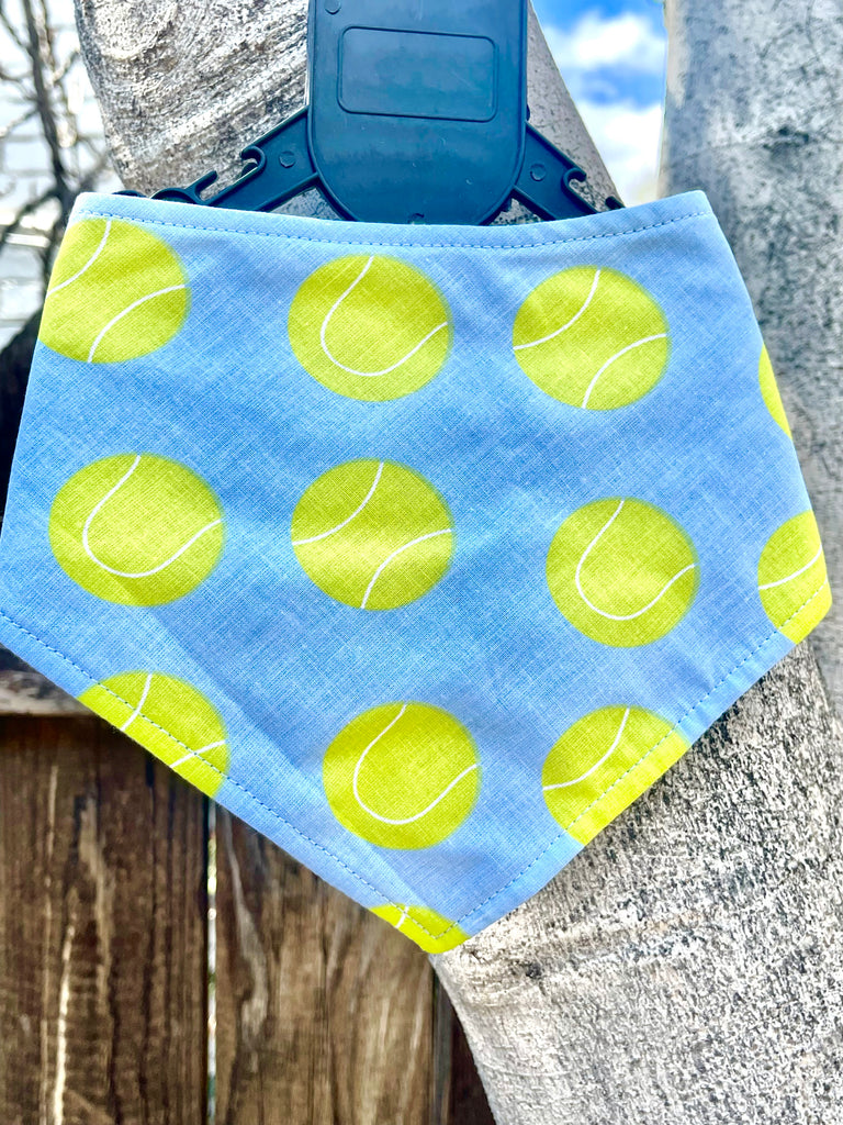 Close-up of a handmade dog bandana featuring a playful tennis ball print in lime green on a light blue-gray background, captured outdoors in natural light.