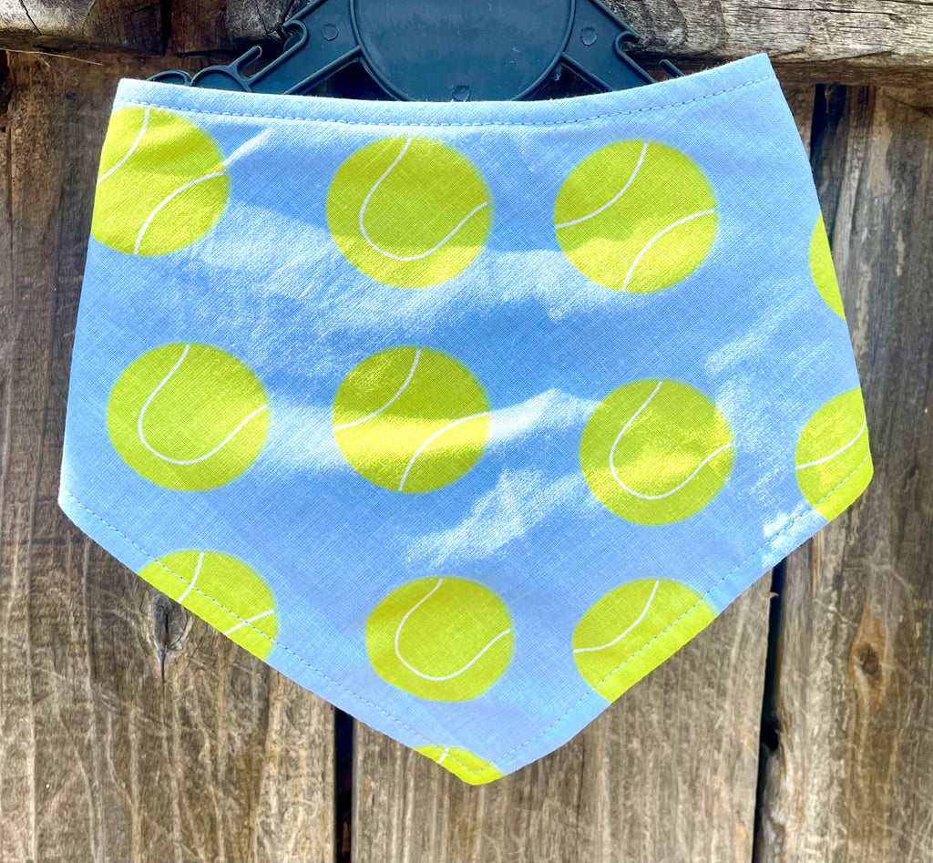 Handmade dog bandana with a light blue-gray background and lime green tennis balls, hanging outdoors in natural sunlight. Perfect for pups that love to play fetch!