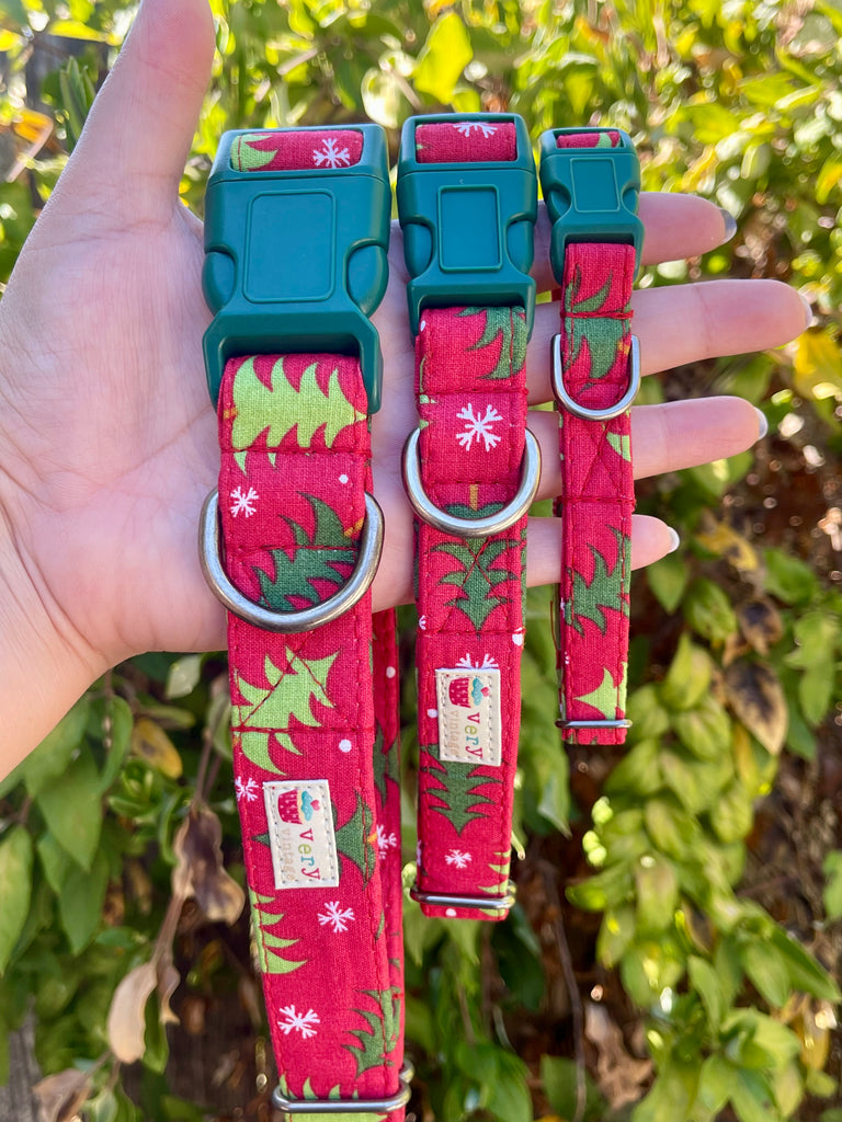 Hand holding three sizes of red collars with green tree shapes and snowflakes, against a leafy green outdoor backdrop. Collars have forest green buckles and silver D-rings.
