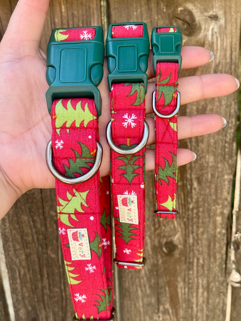 Three sizes of red dog collars with green Christmas tree pattern and white snowflakes, shown against a rustic wood fence. Each collar features a dark green buckle and a metal D-ring.