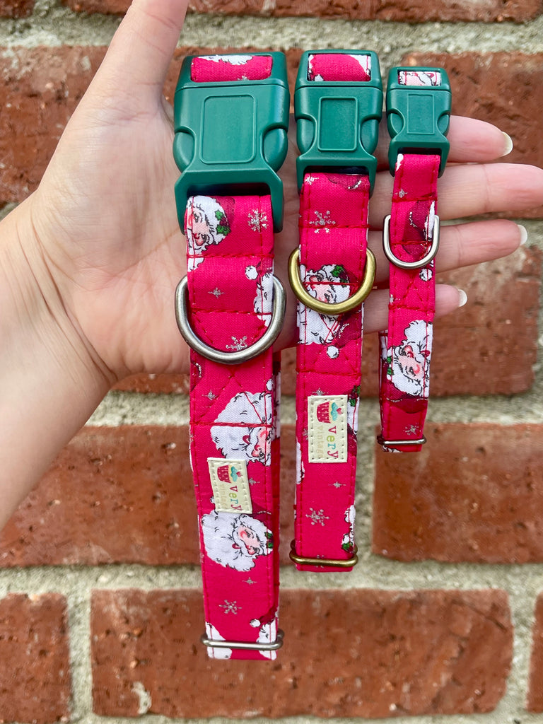 Hand holding three red Santa print dog collars with green buckles against rustic brick wall background