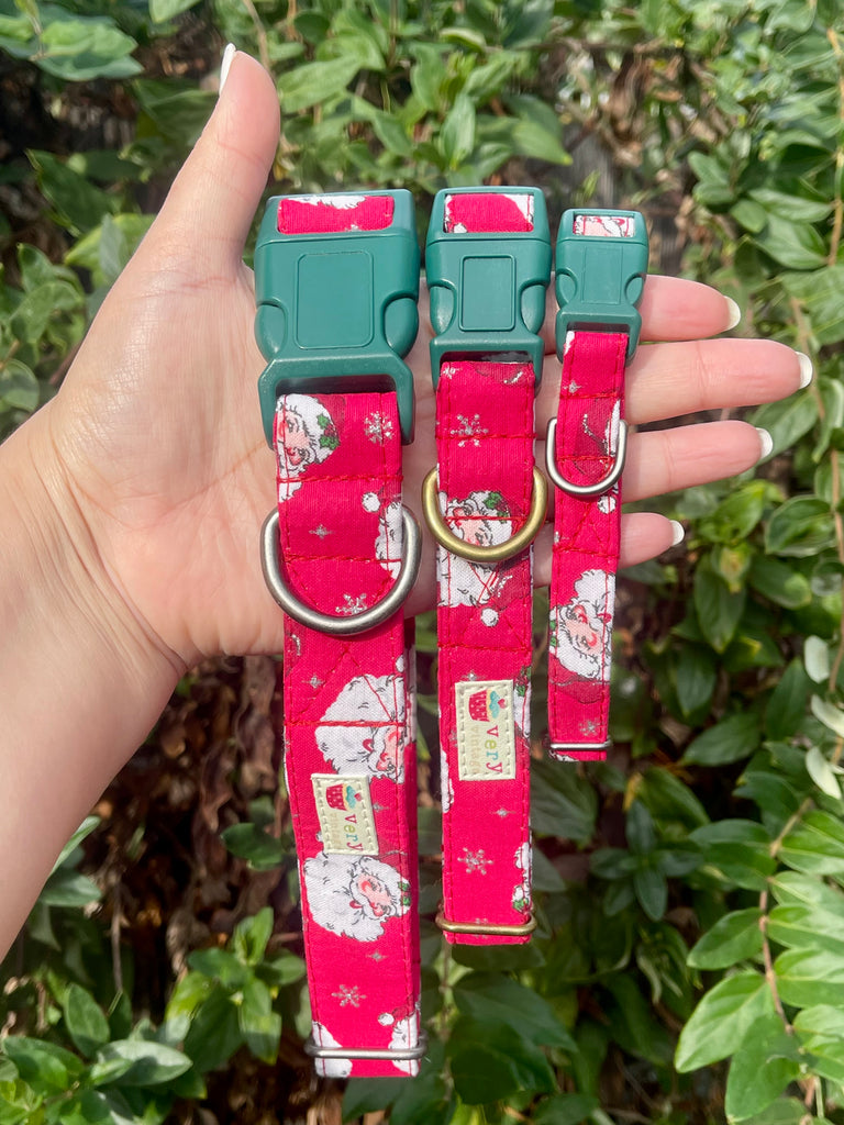 Hand holding three red Santa print collars with green buckles in sunny garden greenery background