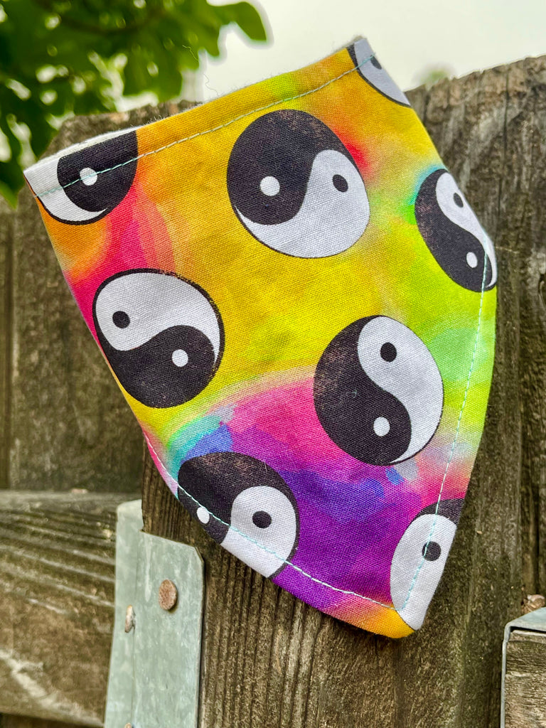 Colorful tie-dye pet bandana with bold black and white yin yang symbols, styled on a weathered wooden fence outdoors.