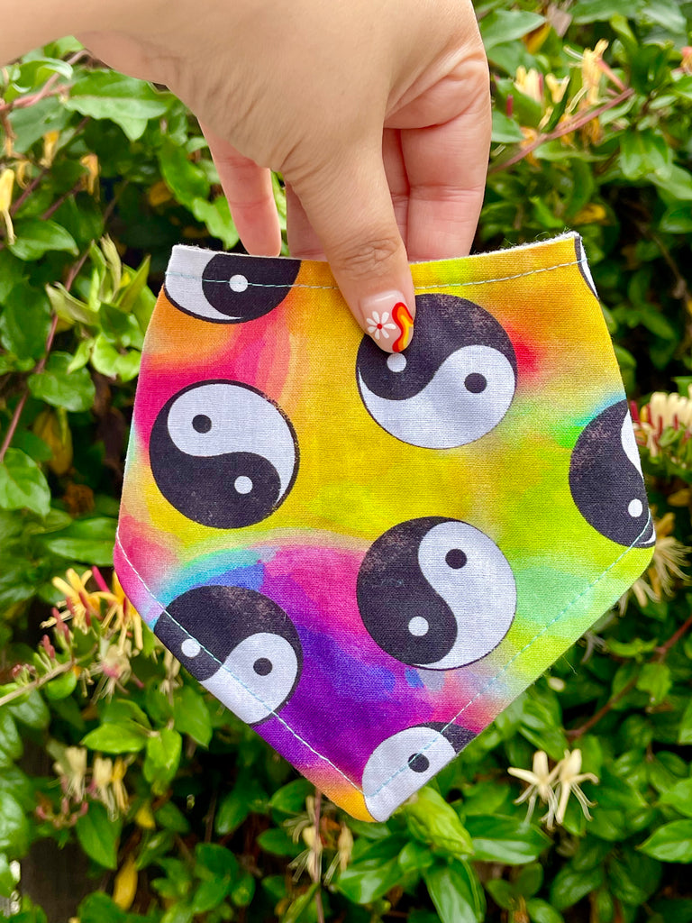 Handmade rainbow tie-dye dog bandana with yin yang design, shown held in hand near blooming garden plants.
